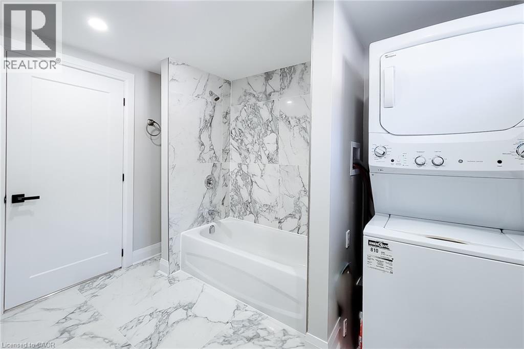 Bathroom featuring a white bathtub with a marble-finish surround, coordinating marble-finish floor tiles, a modern white door with black hardware, and an integrated stacked laundry unit - 103 East 45Th Street, Hamilton, ON - Indoor Photo Showing Laundry Room