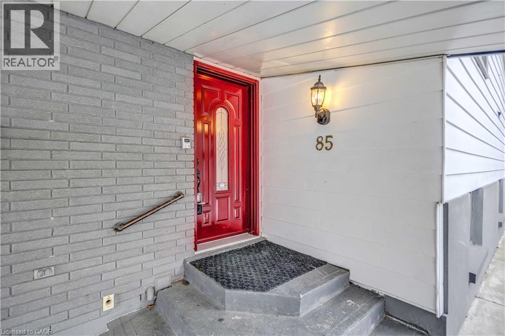 Red entry door with frosted glass panel, flanked by grey brick and white siding - 85 Ranchdale Drive, Hamilton, ON