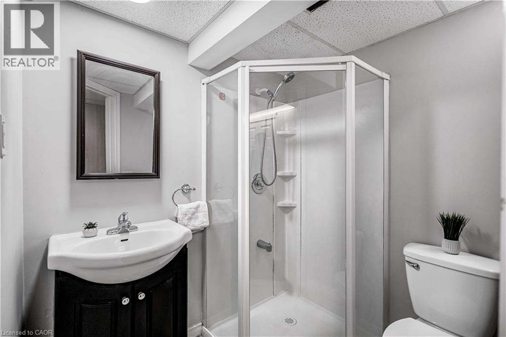 Compact bathroom featuring a corner shower with glass enclosure, pedestal sink with dark wood-finish vanity, wall-mounted mirror, and white toilet - 85 Ranchdale Drive, Hamilton, ON