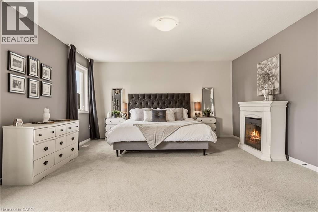 Spacious room featuring light-toned carpeting, a corner fireplace with a white mantel, and a window with dark drapery - 42 Yager Drive, Binbrook, ON - Indoor Photo Showing Bedroom