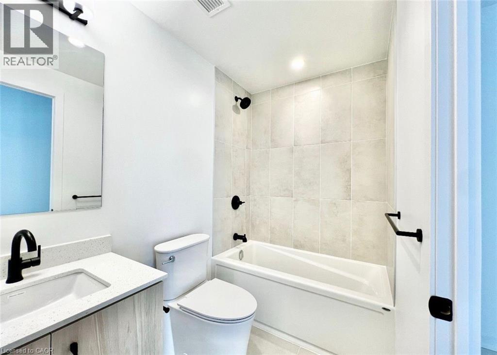 Bathroom featuring a white bathtub with subway tile surround, a light wood-finish vanity with a white countertop and integrated sink, a contemporary black faucet, and a wall-mounted mirror with overhe - 4878 Powers Common Unit# 301, Burlington, ON - Indoor Photo Showing Bathroom