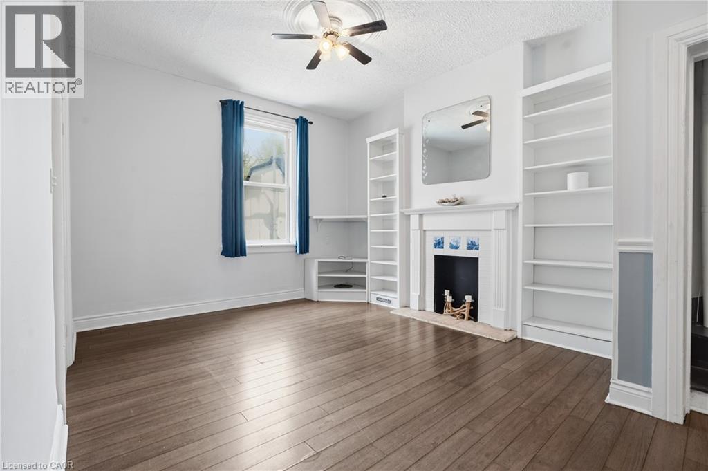 Living area featuring wood-finish flooring, a decorative fireplace with mantel, built-in shelving, a ceiling fan, and a window with natural light - 137 Market Street, Hamilton, ON - Indoor With Fireplace