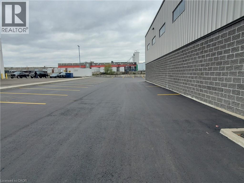 Expansive asphalt parking lot with clearly marked yellow parking lines, featuring a building with a gray brick lower facade and a corrugated metal upper facade with rectangular windows - 44 Ditton Drive Unit# 3, Hamilton, ON