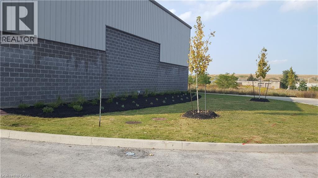 Exterior featuring a gray block wall base, light gray corrugated metal siding, a landscaped planting bed with dark mulch, several young trees, and a concrete curb bordering an asphalt surface - 44 Ditton Drive Unit# 3, Hamilton, ON