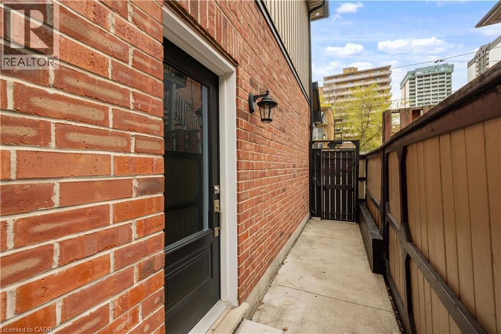 Brick exterior featuring a black entry door with upper glass paneling, an exterior wall lantern, a concrete pathway, and a wooden privacy fence with a gated entry - 199 Hunter Street W, Hamilton, ON - Outdoor With Exterior