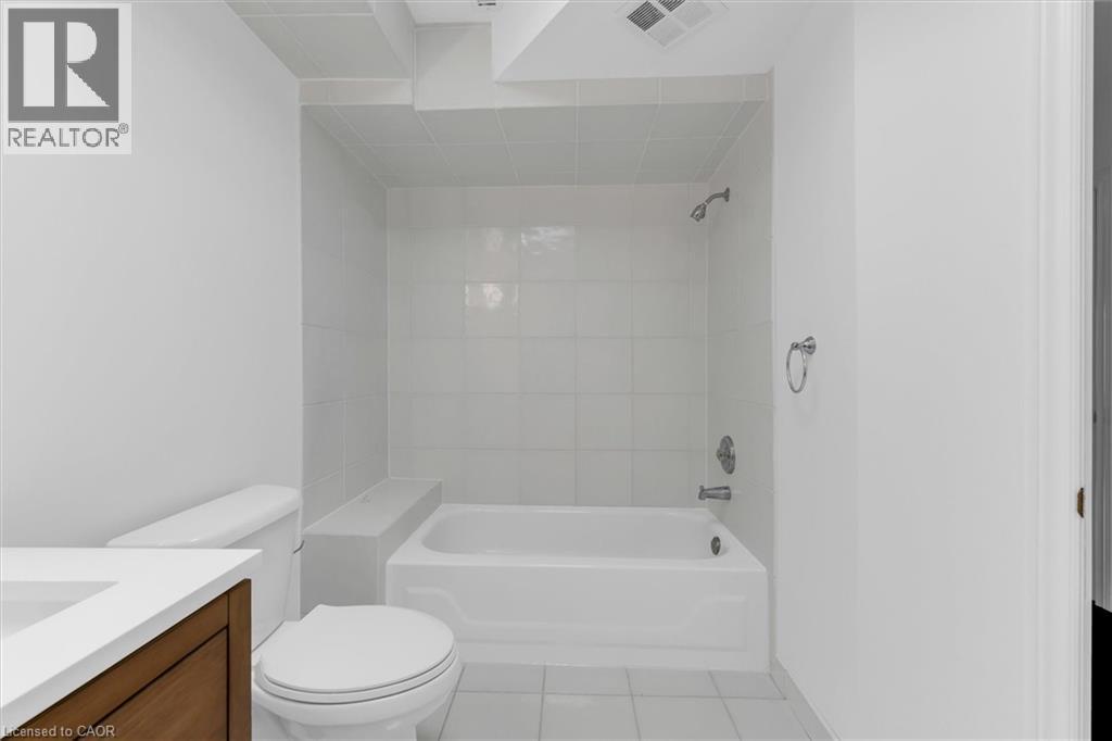 Bathroom featuring a white bathtub with a tiled surround, a toilet, a wood-finish vanity with a white countertop, and tile flooring - 1389 Lakeshore Road, Burlington, ON - Indoor Photo Showing Bathroom
