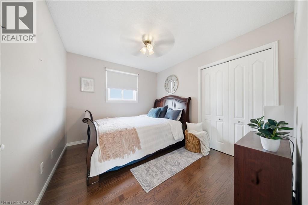 Warm wood-finish flooring throughout, featuring a window with a roller shade, a ceiling fan with light fixture, and white bi-fold closet doors - 200 Morrison Drive, Caledonia, ON - Indoor Photo Showing Bedroom