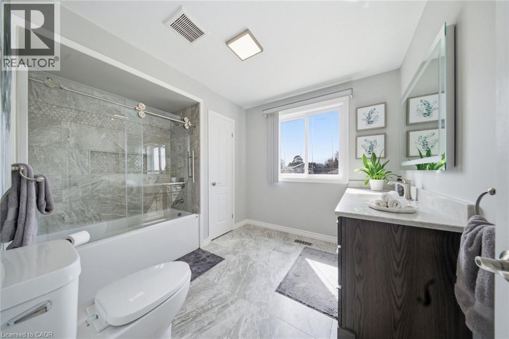 Bathroom featuring a white tub with clear glass sliding doors, gray tiled shower surround, a dark wood-finish vanity with a light countertop and undermount sink, and light gray tile flooring - 200 Morrison Drive, Caledonia, ON - Indoor Photo Showing Bathroom