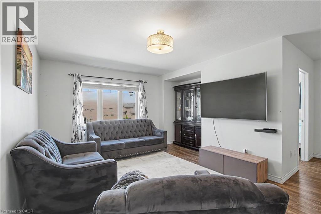 Living area featuring wood-finish flooring, a contemporary ceiling light fixture, a large window with privacy blinds, white painted walls, and a built-in dark wood display cabinet - 60 Postmaster Drive, Kitchener, ON - Indoor Photo Showing Living Room