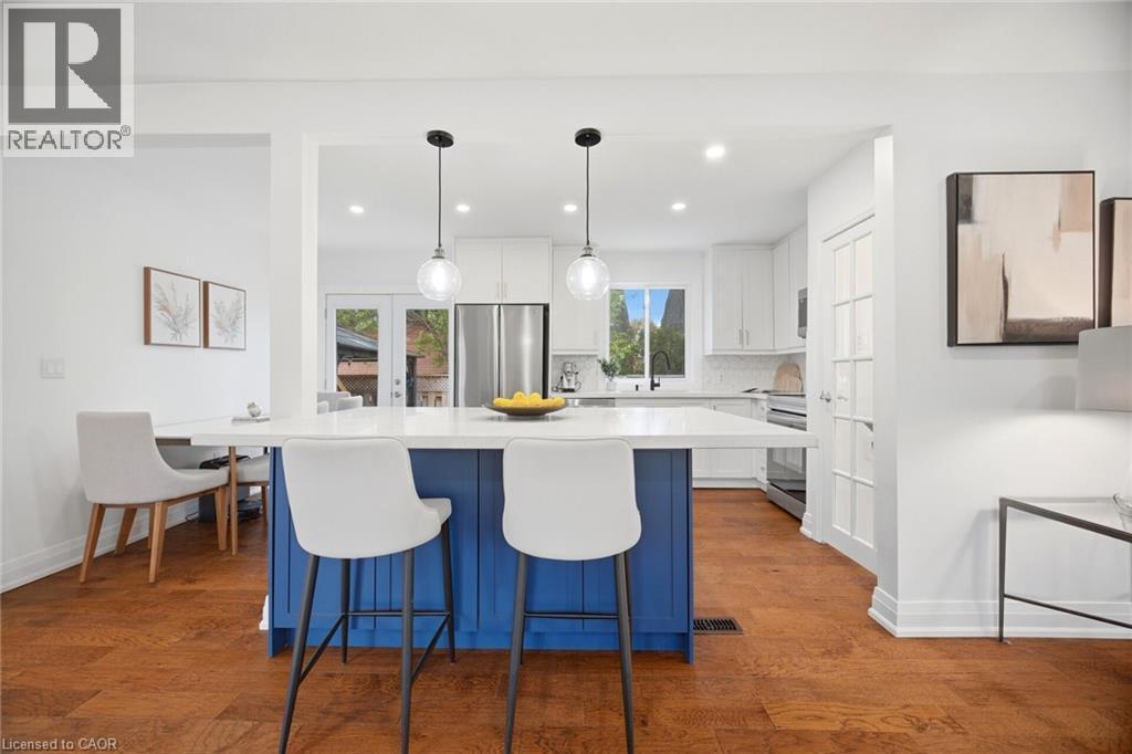 Open-concept kitchen featuring a bold blue island with seating, white cabinetry, integrated stainless steel appliances, and wood-finish flooring - 1376 Royal Drive, Burlington, ON - Indoor