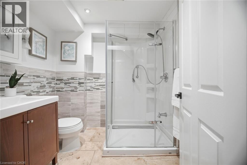 Glass-enclosed shower with multiple spray fixtures, wood-finish vanity with white rectangular sink, white toilet, and neutral-toned floor tiling - 1376 Royal Drive, Burlington, ON - Indoor Photo Showing Bathroom