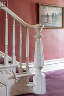 Ornate white wooden newel post and balusters with carved detailing, framing a staircase featuring purple patterned carpeting -