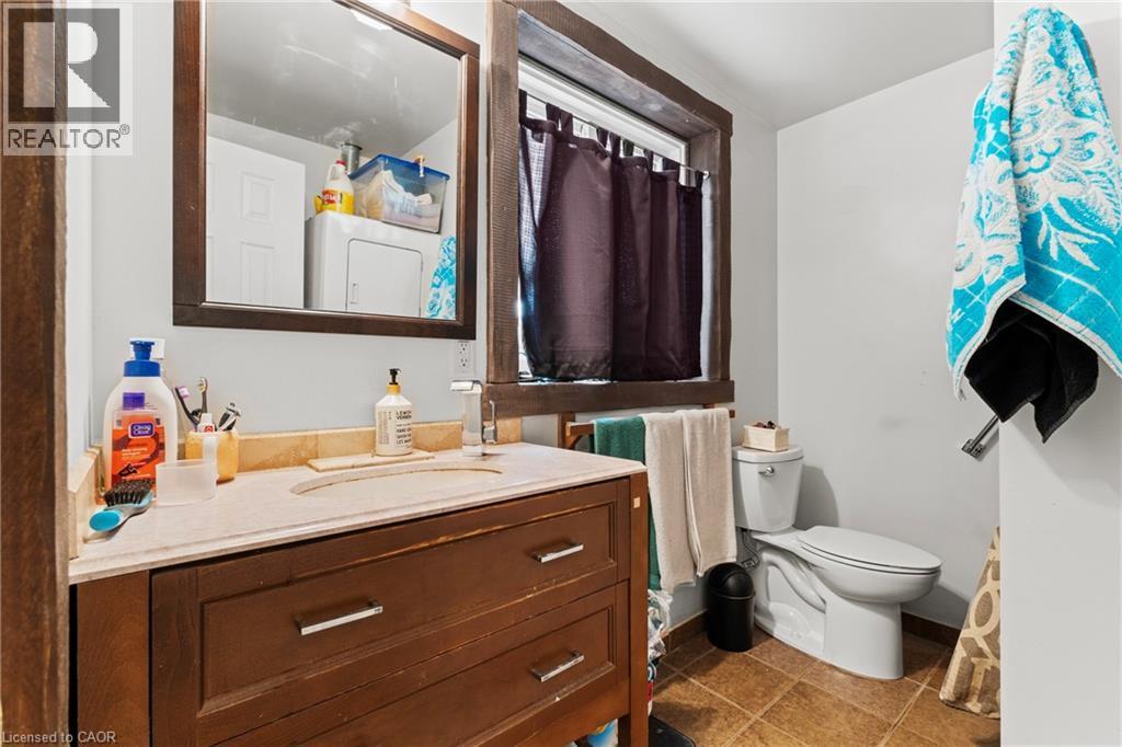 Bathroom featuring a wood-finish vanity with an integrated sink and light-toned countertop, framed mirror, and a window with dark trim - 3 San Pedro Drive, Hamilton, ON - Indoor Photo Showing Bathroom