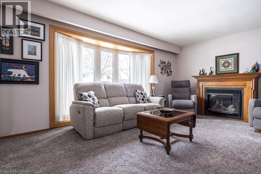 Living area featuring a prominent wood fireplace with a detailed mantel, a bay window with a wood-finish frame, light-toned wall paint, and gray carpet flooring - 19 Chateau Court, Hamilton, ON - Indoor Photo Showing Living Room With Fireplace