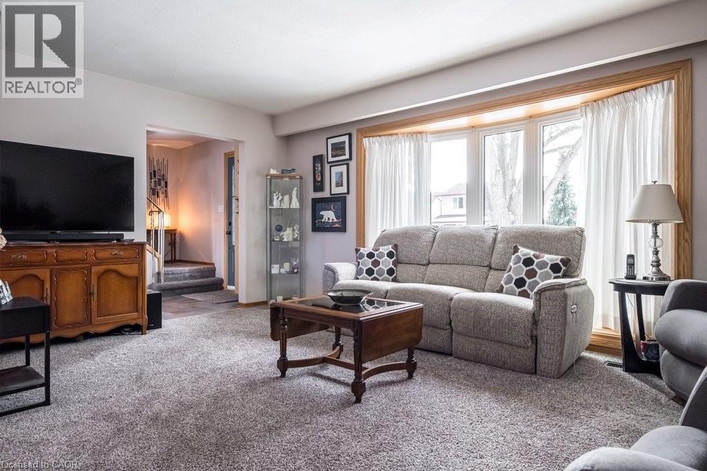 Living area featuring a bay window with wood trim, gray carpeting, neutral wall colors, and a partial view of a staircase - 19 Chateau Court, Hamilton, ON - Indoor Photo Showing Living Room
