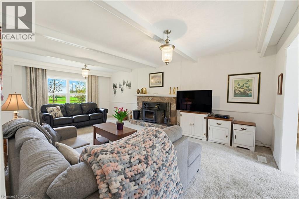 Living area featuring a stone fireplace with a wood stove insert, exposed ceiling beams, and light-colored carpet flooring - 10 Huisman Lane, Hamilton, ON