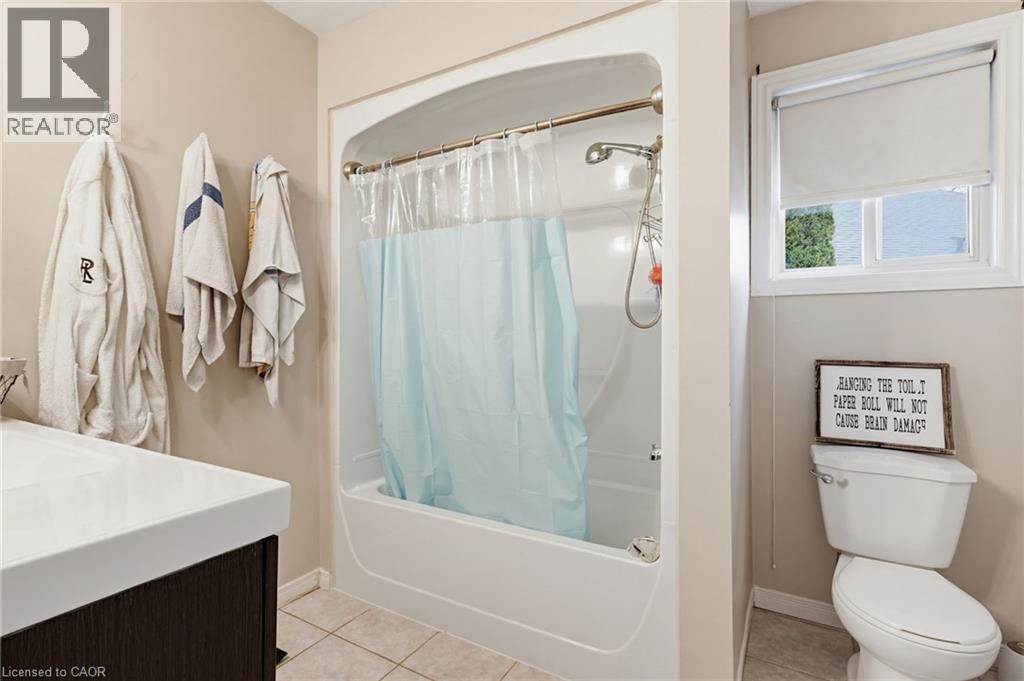 Bathroom featuring a tub/shower combination, a dark wood-finish vanity with an integrated white sink, a window with a roller blind, and light-toned floor tiles - 47 Hannah Crescent, Hamilton, ON - Indoor Photo Showing Bathroom