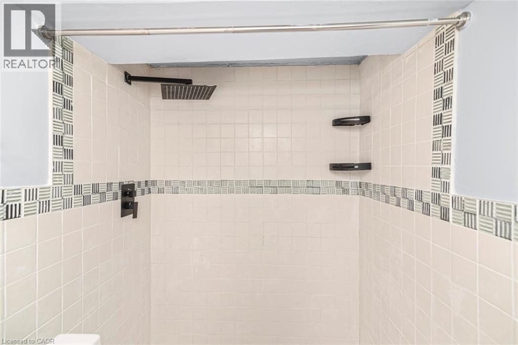 Shower enclosure featuring cream-colored square tiles, a decorative mosaic tile accent band, a square matte black rainfall showerhead, and matte black corner shelving - 112 Nash Road S, Hamilton, ON - Indoor Photo Showing Bathroom