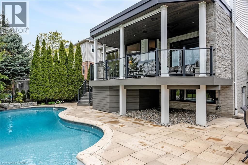 Exterior featuring a freeform swimming pool, stone patio, and an elevated covered deck with glass railings - 4285 Couples Crescent, Burlington, ON - Outdoor With In Ground Pool With Deck Patio Veranda