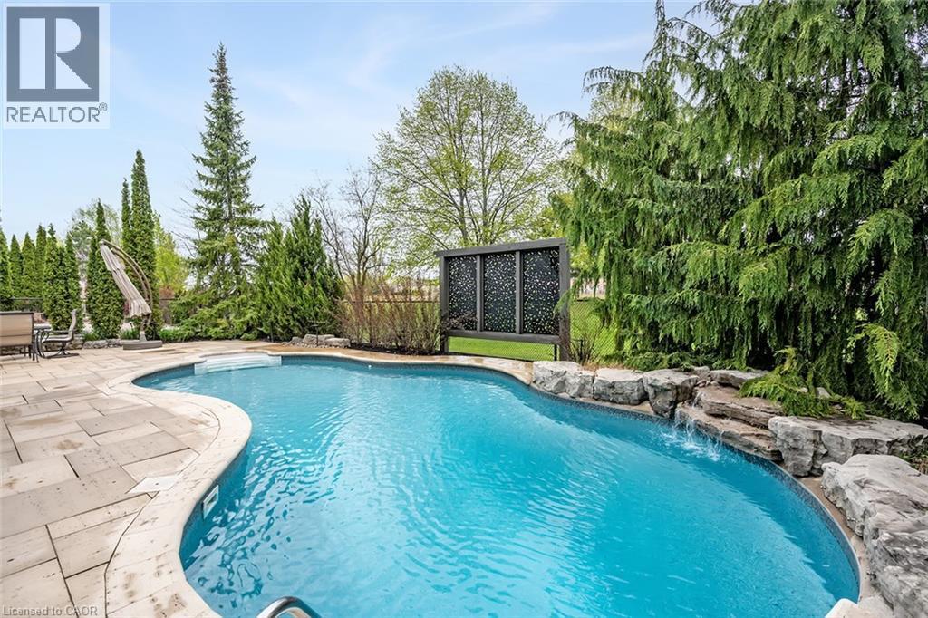 Curved inground pool featuring a stone waterfall, a surrounding paver patio, and lush evergreen landscaping - 4285 Couples Crescent, Burlington, ON - Outdoor With In Ground Pool With Backyard