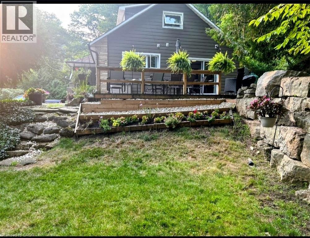 Rear exterior featuring a spacious wooden deck with railings, stone retaining walls, terraced gardens, and sliding glass doors - 1333 Hidden Valley Road, Burlington, ON - Outdoor With Deck Patio Veranda