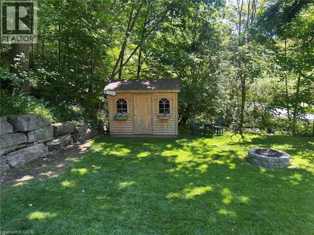 Expansive grassy yard featuring a wood-sided shed with arched windows, a stone block retaining wall, and a built-in stone fire pit - 1333 Hidden Valley Road, Burlington, ON - Outdoor