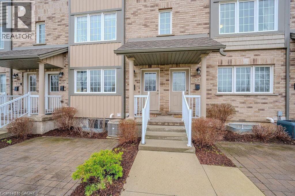 Brick townhouse exterior featuring multiple white-framed windows, a covered entrance with white railings, and a paver stone patio area - 275 Old Huron Road Unit# 21, Kitchener, ON - Outdoor With Facade