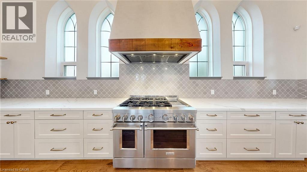 Gourmet kitchen featuring a professional-grade stainless steel range, custom range hood with wood accent, white shaker cabinetry with dark hardware, white countertops, and arabesque tile backsplash - 41 Talbot Road, Delhi, ON - Indoor Photo Showing Kitchen With Upgraded Kitchen