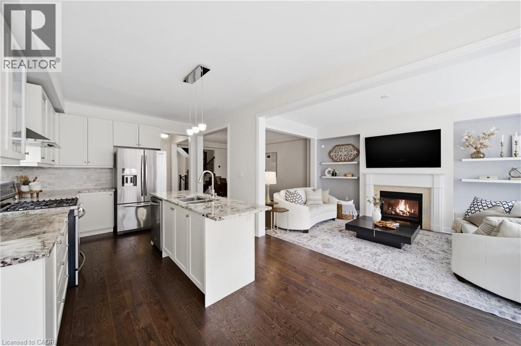 Open-concept kitchen featuring white cabinetry, stainless steel appliances, a center island with sink, and granite countertops - 2466 Village Common, Oakville, ON - Indoor Photo Showing Other Room With Fireplace
