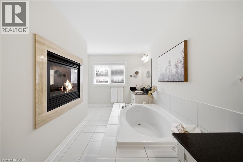 Recessed fireplace with a stone surround, integrated soaking tub, white tile flooring, double vanity, and a window with blinds - 2466 Village Common, Oakville, ON - Indoor Photo Showing Bathroom