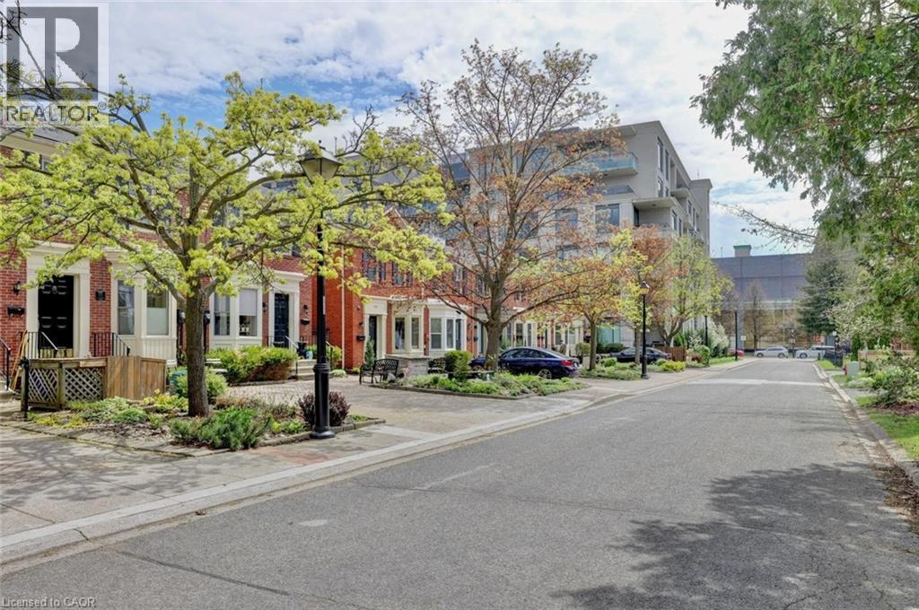 Brick row homes featuring dark entry doors with white trim, front landscaping, and mature trees lining the street - 463 Blathwayte Lane, Burlington, ON - Outdoor With Facade