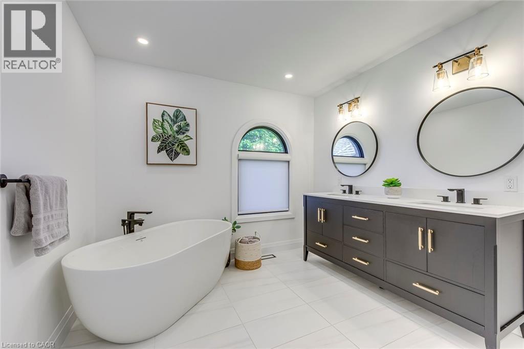 White freestanding soaking tub paired with a double vanity featuring a dark finish, gold hardware, and two circular framed mirrors - 54 Deerview Avenue, Ancaster, ON - Indoor Photo Showing Bathroom