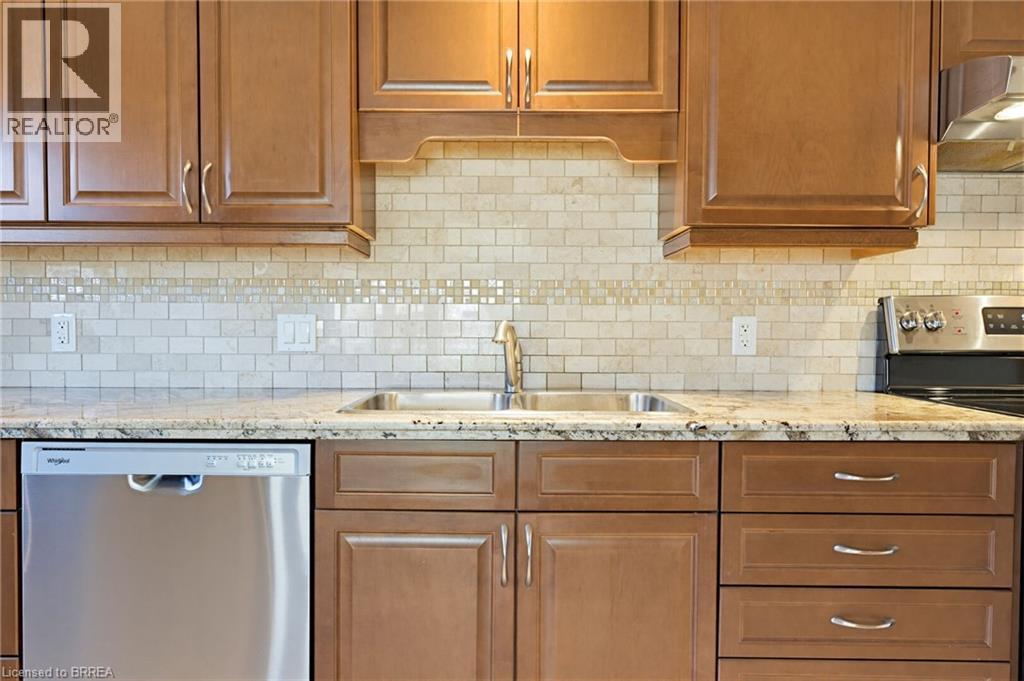 Kitchen featuring warm-toned cabinetry, a subway tile backsplash with decorative accents, light-colored stone countertops, and a double basin stainless steel sink - 6 General Street, Brantford, ON - Indoor Photo Showing Kitchen With Double Sink