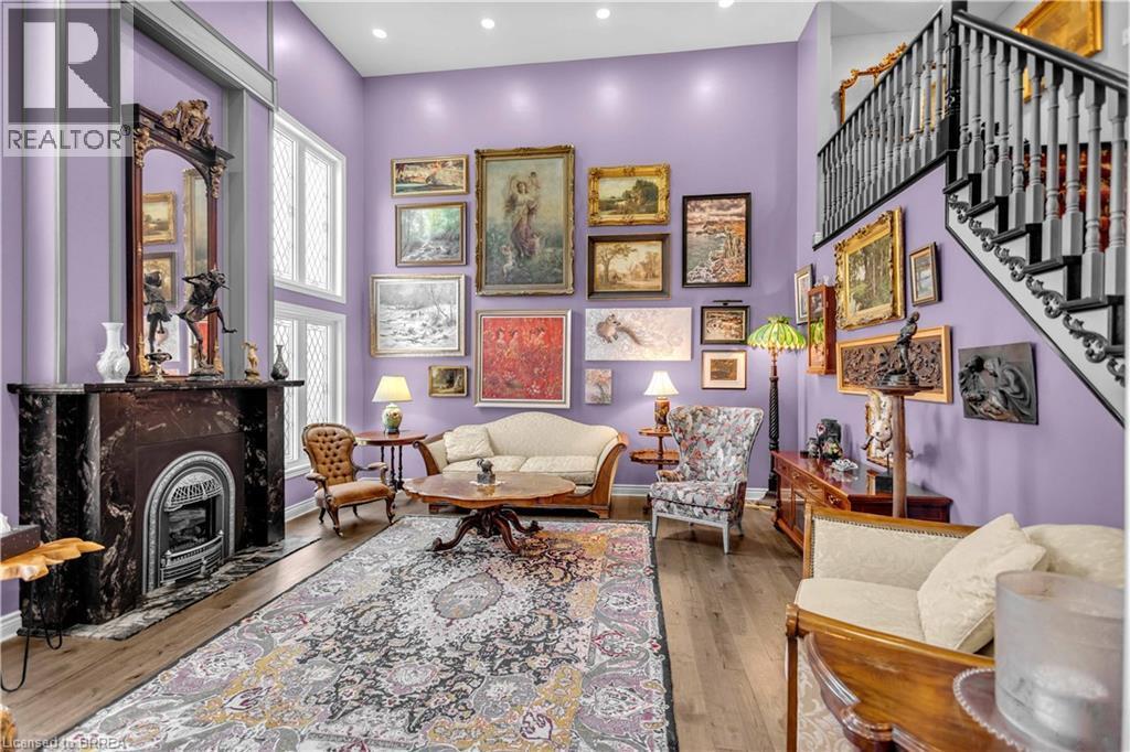 Double-height living space featuring a dark stone fireplace with decorative mantel, wood-finish flooring, a grand staircase with dark railings, and ample natural light from tall windows - 69 Hunter Way, Brantford, ON - Indoor
