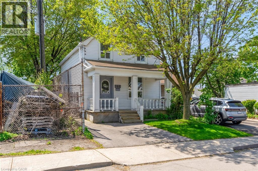 White siding residence featuring a covered front porch with white railing, a mature front yard tree, a paved driveway, and a chain-link fence - 168 Britannia Avenue, Hamilton, ON - Outdoor With Deck Patio Veranda