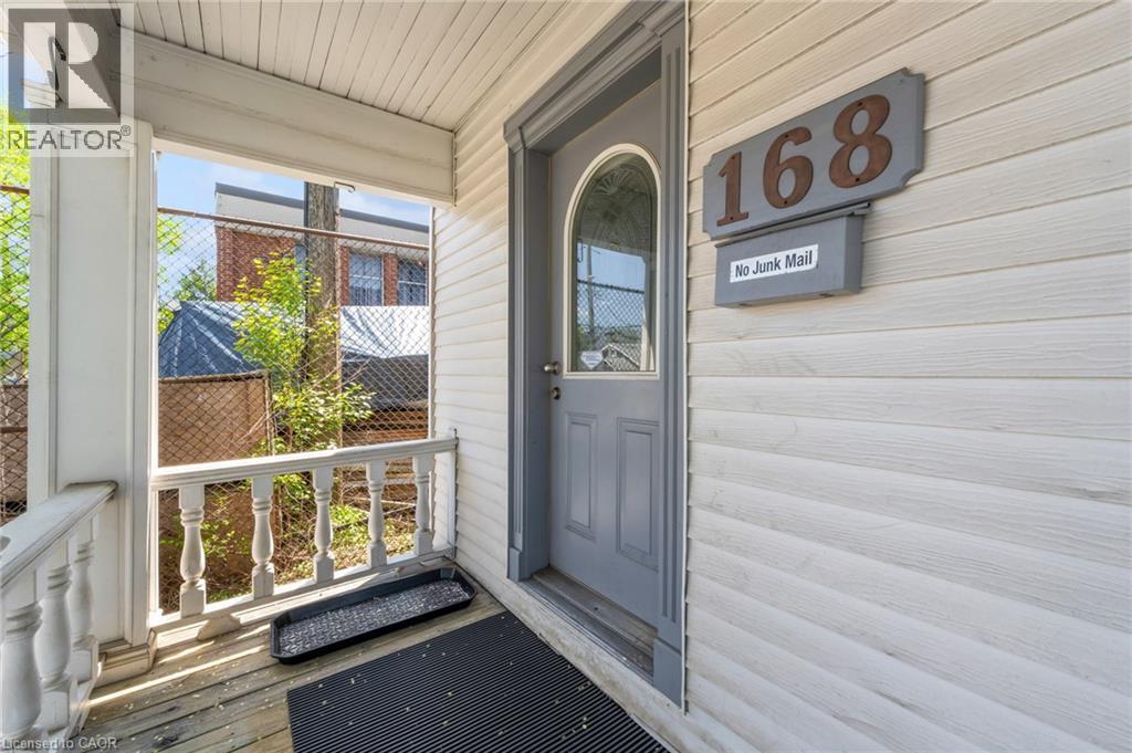 Front entry featuring a gray paneled door with an arched glass insert, white horizontal siding, and a covered porch with white turned balusters - 168 Britannia Avenue, Hamilton, ON - Outdoor With Exterior