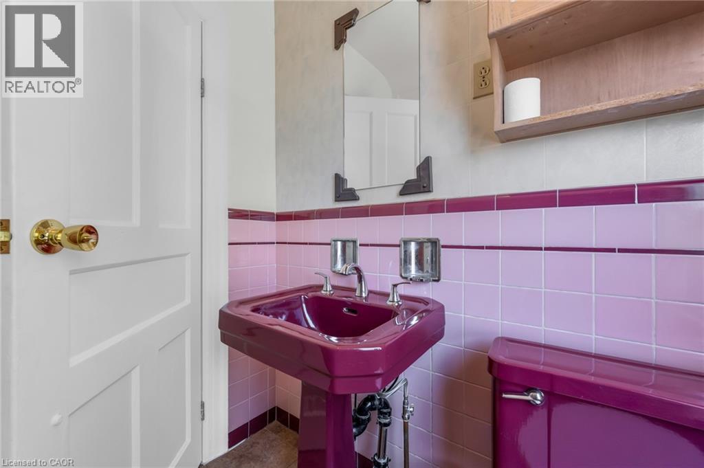 Pedestal sink with coordinating toilet, pink and purple tiled walls, polished brass hardware, and an overhead wooden shelf - 168 Britannia Avenue, Hamilton, ON - Indoor
