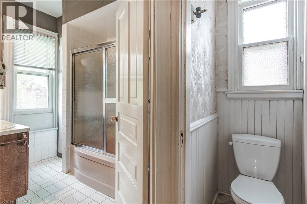 Bathroom featuring a white tile floor, a window with blinds, a shower-over-tub with a sliding glass door, a wood-finish vanity with a white countertop, and white beadboard paneling - 18 Fairleigh Crescent, Hamilton, ON - Indoor Photo Showing Bathroom