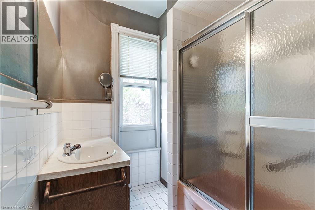 Bathroom featuring a wood-finish vanity with an integrated sink and chrome faucet, white tiled walls and flooring, and a shower with sliding frosted glass doors - 18 Fairleigh Crescent, Hamilton, ON - Indoor Photo Showing Bathroom