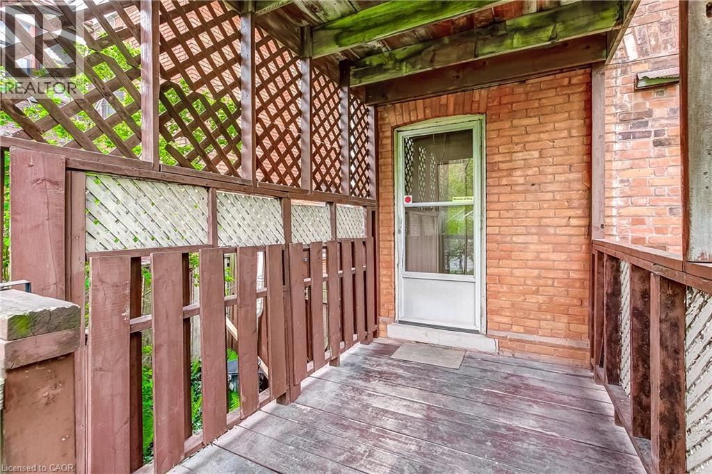 Covered wood-finish porch featuring a white screen door, brick exterior wall, wood railing, and decorative lattice panels - 18 Fairleigh Crescent, Hamilton, ON - Outdoor With Exterior