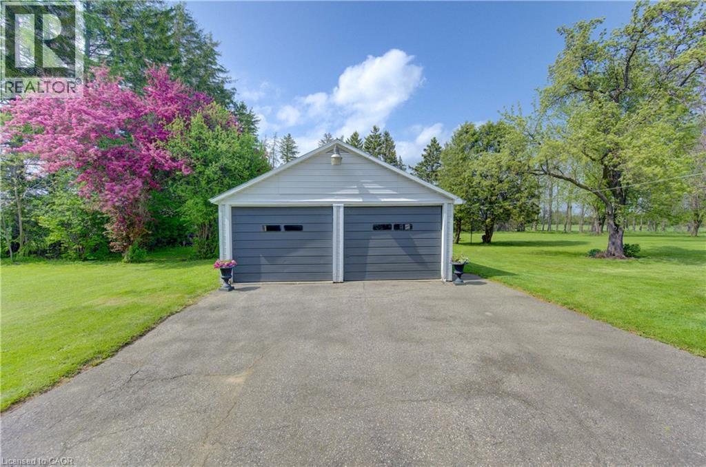 Detached two-car garage with light gray siding and dark gray garage doors, set on a paved driveway with surrounding green lawn and mature trees - 1463 Kossuth Road, Cambridge, ON - Outdoor