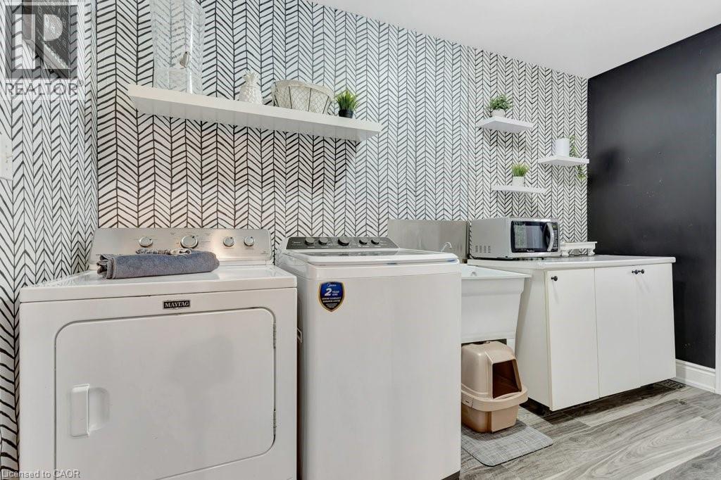 Functional laundry area featuring graphic pattern wallpaper, wood-finish flooring, white cabinetry, a utility sink, and floating shelves - 49 Small Court, Cambridge, ON - Indoor Photo Showing Laundry Room