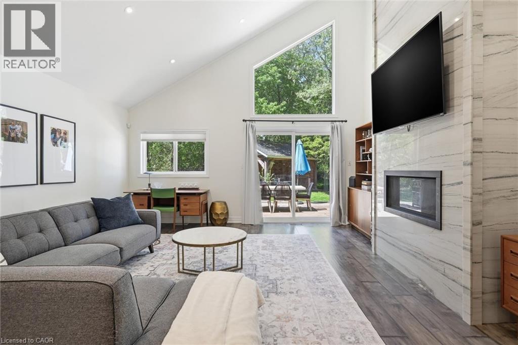 Vaulted ceiling living area featuring wood-finish flooring, a contemporary fireplace with a stone surround, and abundant natural light from multiple windows, including a large triangular window - 24 Glenwood Crescent, Hamilton, ON - Indoor Photo Showing Living Room With Fireplace