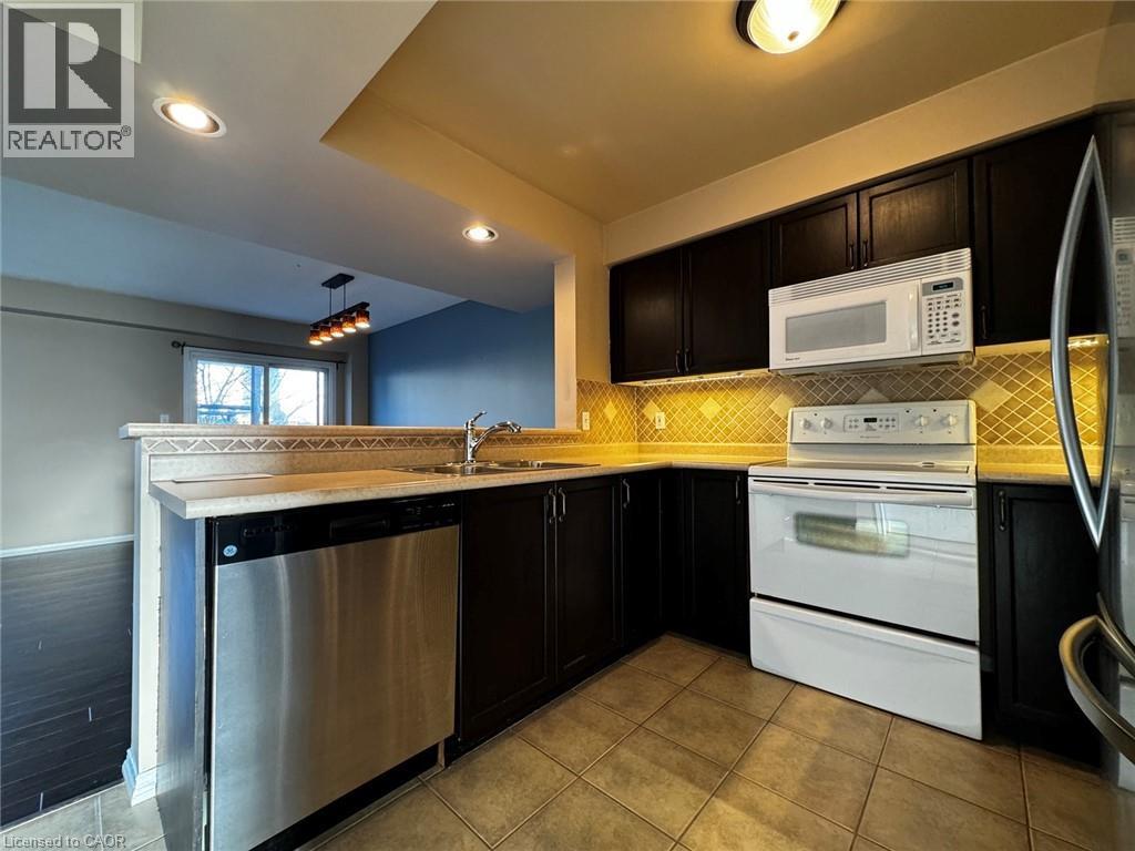 Kitchen featuring dark cabinetry, stainless steel dishwasher, white range with overhead microwave, tan tile flooring, and decorative tile backsplash - 37 Northcliffe Crescent, Cambridge, ON - Indoor Photo Showing Kitchen With Double Sink