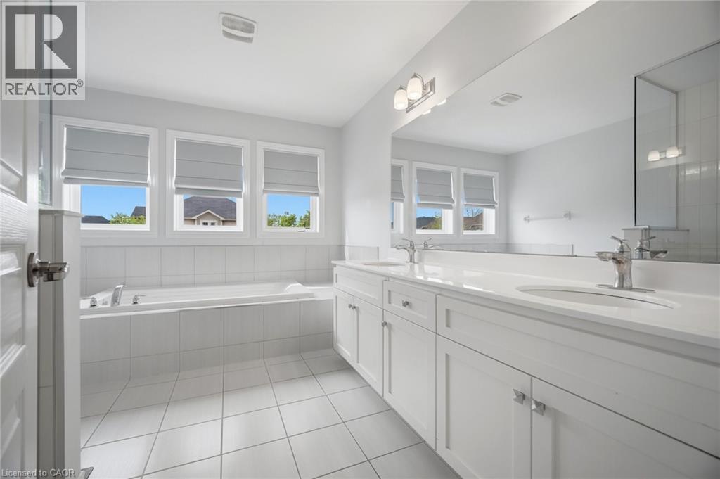 Bathroom featuring a double vanity with white cabinetry and integrated sinks, a built-in bathtub with tiled surround, three windows with blinds, and white square floor tiles - 58 Doon Creek Street, Kitchener, ON - Indoor Photo Showing Bathroom