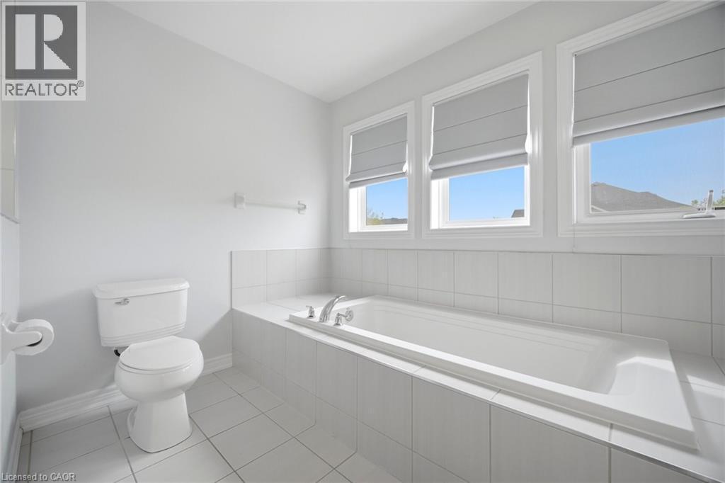 Bathroom featuring a built-in tub with chrome fixtures, three windows with privacy shades, and white tile surround - 58 Doon Creek Street, Kitchener, ON - Indoor Photo Showing Bathroom