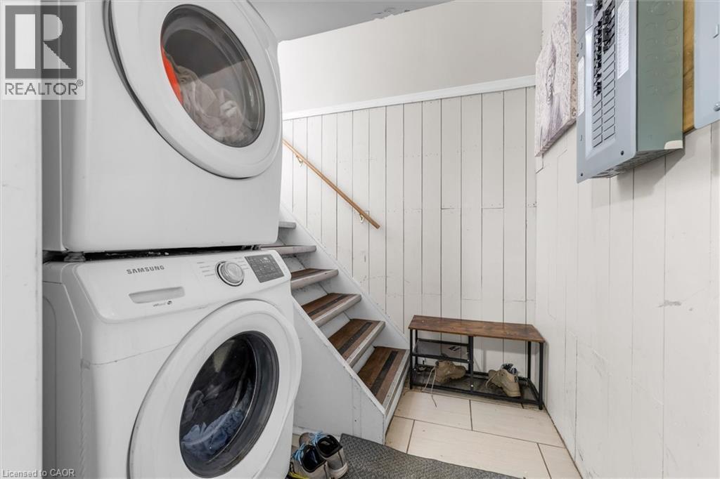 Dedicated laundry area featuring a stacked washer and dryer, white paneled walls, a neutral-toned tiled floor, and a wooden handrail along the adjacent staircase - 46 Beaconsfield Drive, Hamilton, ON - Indoor Photo Showing Laundry Room