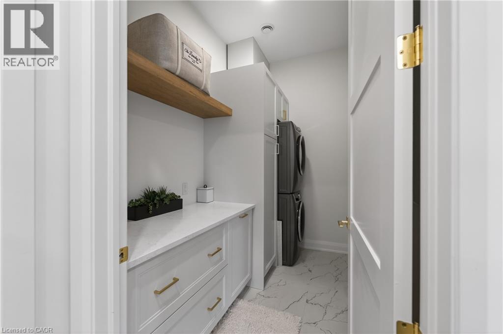 Dedicated laundry area featuring a stacked washer and dryer, white cabinetry with gold-tone hardware, a light-toned countertop, a floating wood shelf, and marble-look floor tiles - 2 Arbourvale Common Unit# 410, St. Catharines, ON - Indoor