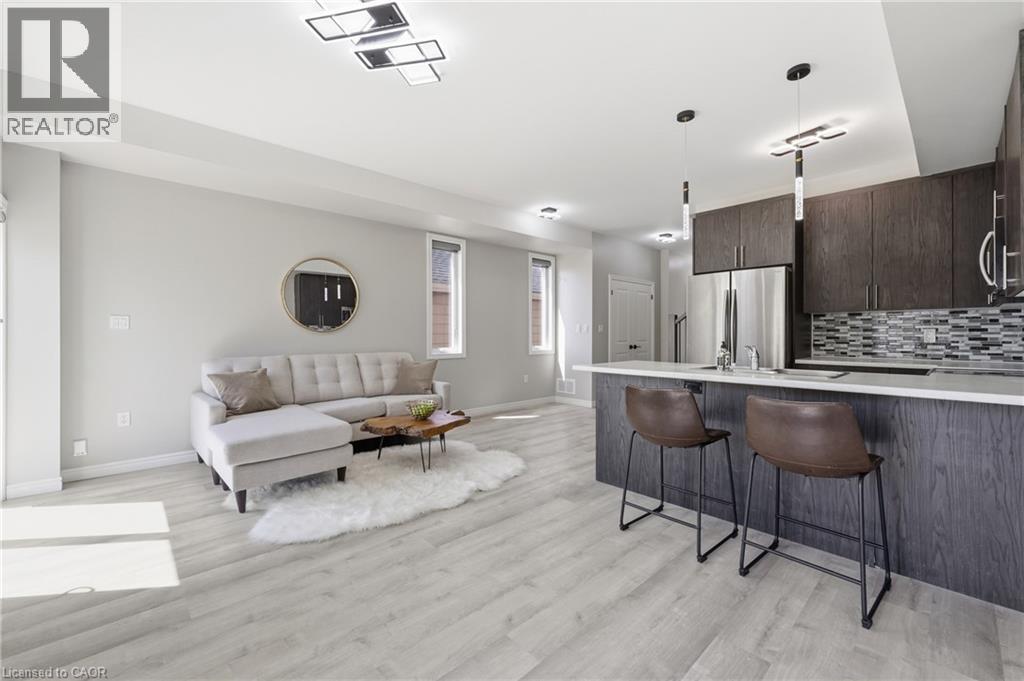 Open-concept living space featuring light wood-finish flooring, a contemporary kitchen island with a white countertop, dark wood-finish cabinetry, stainless steel appliances, and a mosaic tile backspl - 37 Delena Avenue S, Hamilton, ON - Indoor Photo Showing Kitchen With Upgraded Kitchen