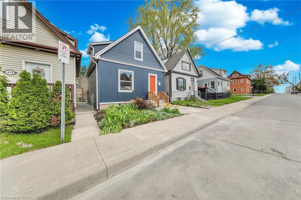 Blue vertical siding exterior featuring a contrasting orange front door, a concrete sidewalk, and a landscaped front yard with varied plantings - 501 Ferguson Avenue N, Hamilton, ON - Outdoor With Facade